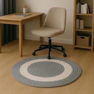 May include: A grey and white round braided rug with a white border. The rug is placed under a beige office chair with a wooden desk in the background.