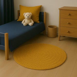 May include: A round, yellow, crocheted rug with a braided texture. The rug is on a wooden floor in a room with a blue bed and a wooden dresser.