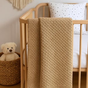 Peut inclure: Une couverture bébé en crochet beige drapé sur un lit bébé en bois avec un oreiller blanc à pois noirs.