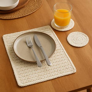 Può includere: Un sottopentola in crochet bianco con un piatto marrone, una forchetta e un coltello sopra. Ci sono due sottobicchieri in crochet bianco, uno con un bicchiere di succo d'arancia e l'altro vuoto.