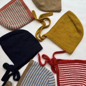 以下が含まれることがあります： 様々な色のニットベビーボンネットのコレクション。錆色、マスタードイエロー、ネイビーブルー、ブラウンなど。ストライプ模様のものもあれば、無地のものもあります。各ボンネットには結び紐が付いています。