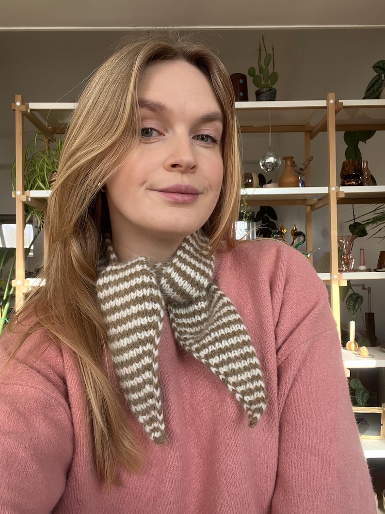 May include: A knitted scarf with alternating stripes of white and brown. The scarf is tied around the neck, with the ends hanging down. The person is wearing a pink sweater. Shelves with plants are in the background.