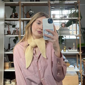 May include: A light yellow knitted scarf is worn over a pink button-down shirt. The scarf is tied in a knot. The shirt has long sleeves and a chest pocket. The background includes a wooden shelf with plants and decorative items.