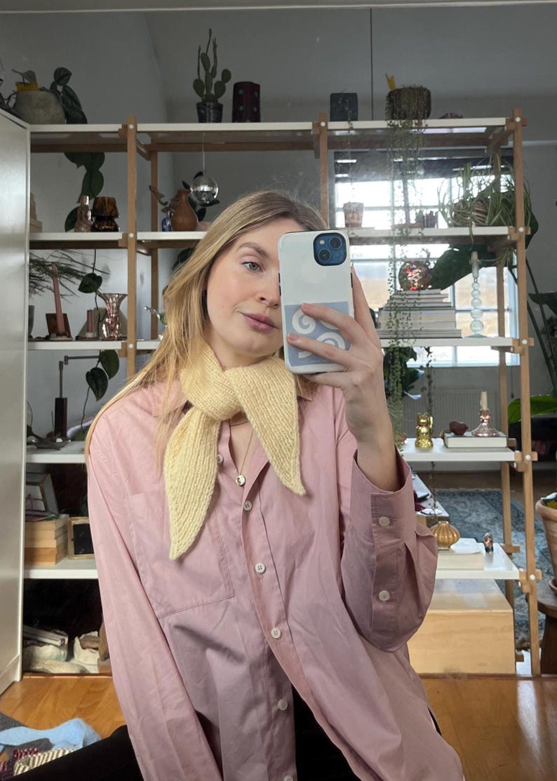 May include: A light yellow knitted scarf is worn over a pink button-down shirt. The scarf is tied in a knot. The shirt has long sleeves and a chest pocket. The background includes a wooden shelf with plants and decorative items.