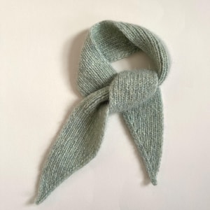 May include: A light blue knitted scarf, knotted in the center. The scarf has a soft, textured appearance and is made of a light, airy material. The ends of the scarf are pointed. The scarf is displayed on a white background.