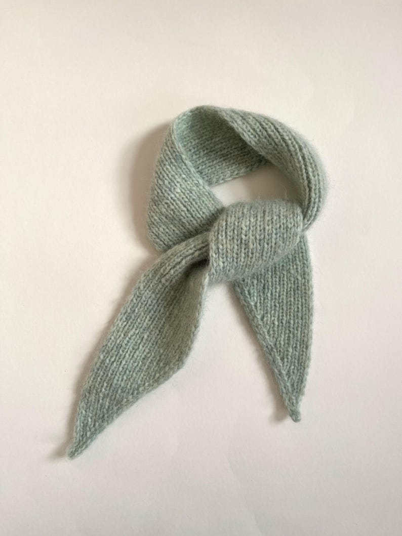 May include: A light blue knitted scarf, knotted in the center. The scarf has a soft, textured appearance and is made of a light, airy material. The ends of the scarf are pointed. The scarf is displayed on a white background.