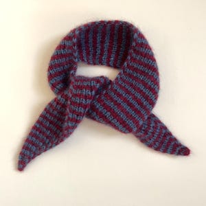 May include: A knitted scarf in a bandana style, featuring alternating stripes of burgundy and blue. The scarf is folded and tied, with the ends pointing downwards. The material appears soft and textured, suitable for cold weather.