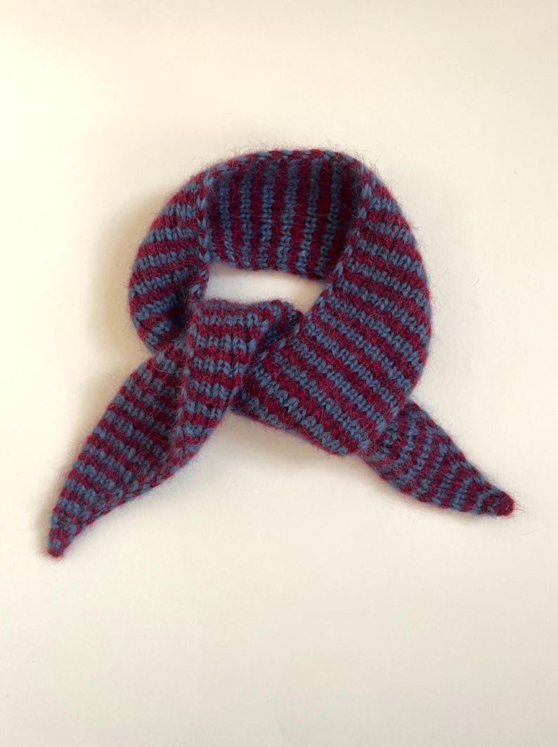 May include: A knitted scarf in a bandana style, featuring alternating stripes of burgundy and blue. The scarf is folded and tied, with the ends pointing downwards. The material appears soft and textured, suitable for cold weather.