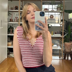 Könnte beinhalten: Ein ärmelloses Häkeltop mit V-Ausschnitt in einem rot-weiß karierten Muster. Das Top wird mit einer dunkelblauen Hose kombiniert. Das Bild wird in Innenräumen aufgenommen, wobei eine Person ein Telefon vor sich hält.