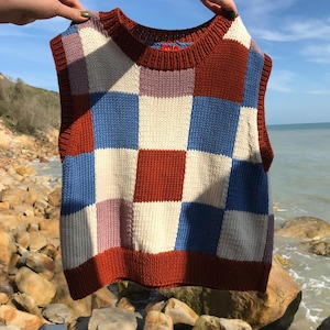 Peut inclure: Un gilet tricoté aux motifs patchwork. Il présente des carrés de différentes couleurs, notamment du rouge, du bleu, du blanc et du rose. Le vêtement sans manches est présenté devant un ciel bleu et la mer.