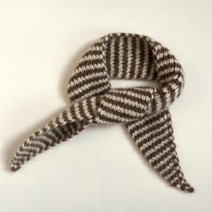 May include: A brown and white striped knitted scarf, knotted in the center. The scarf is made of a soft, textured yarn and has a simple, classic design. The scarf is displayed on a white background.