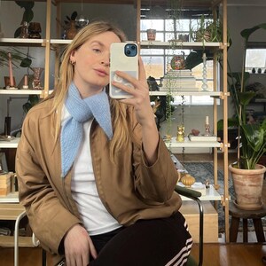 May include: A light blue knit scarf with a pointed end is worn over a white shirt and a brown jacket. The person is wearing black sweatpants with pink stripes. The background includes a wooden shelf with plants and other decorative items.