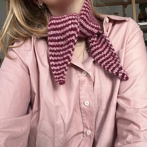 May include: A knitted scarf in shades of pink and burgundy, featuring horizontal stripes. The scarf is tied around the neck, with the ends hanging down. The person is wearing a pink button-down shirt.