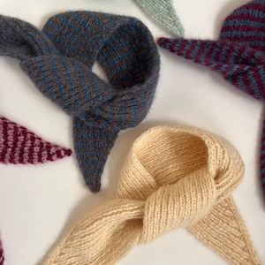 May include: A collection of knitted neck scarves in various colors. The scarves are in shades of blue, brown, burgundy, and cream. Each scarf is knotted at the center and has pointed ends. The items are arranged on a white surface.