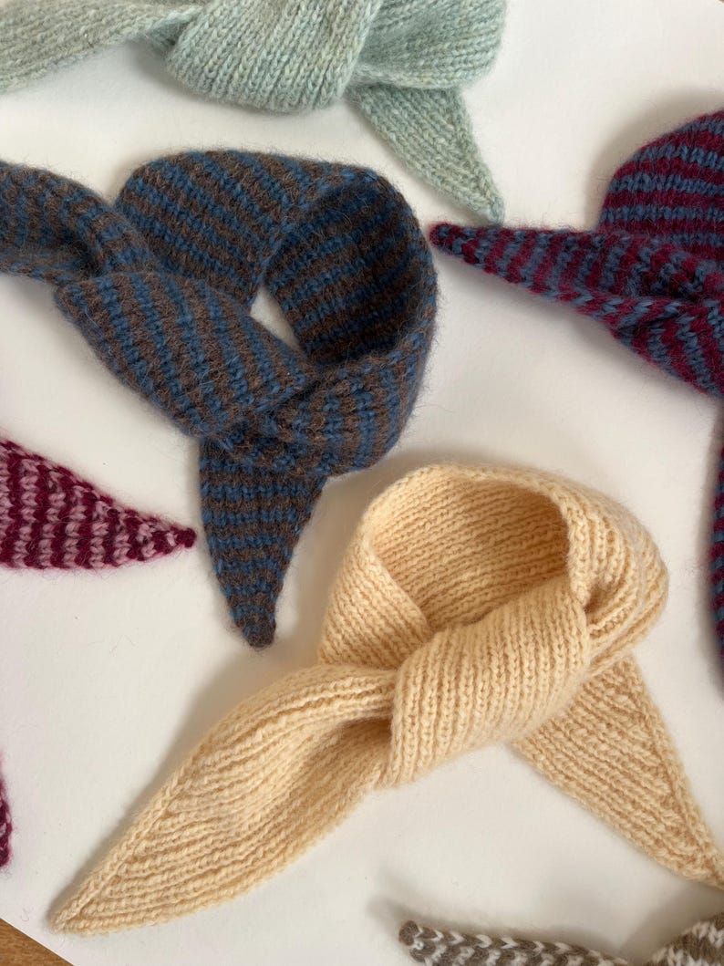 May include: A collection of knitted neck scarves in various colors. The scarves are in shades of blue, brown, burgundy, and cream. Each scarf is knotted at the center and has pointed ends. The items are arranged on a white surface.