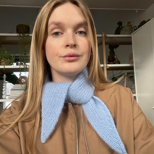 May include: A light blue knitted scarf with pointed ends. The scarf is worn over a brown jacket with a zipper. The scarf is made of a soft, textured yarn.