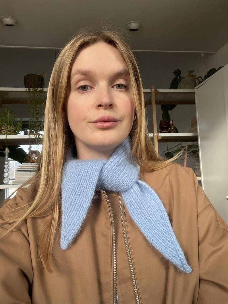 May include: A light blue knitted scarf with pointed ends. The scarf is worn over a brown jacket with a zipper. The scarf is made of a soft, textured yarn.