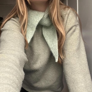 May include: A light green knit scarf is worn over a light green sweater. The scarf is triangular and has a ribbed texture. The sweater has a soft, fuzzy texture. The person's blonde hair is visible.