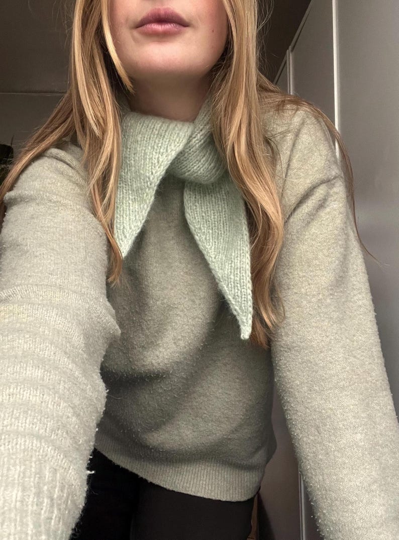 May include: A light green knit scarf is worn over a light green sweater. The scarf is triangular and has a ribbed texture. The sweater has a soft, fuzzy texture. The person's blonde hair is visible.