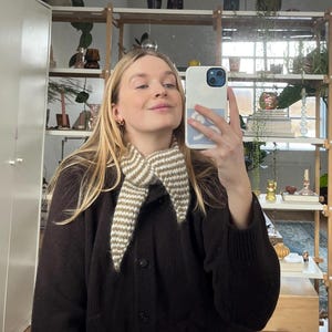 May include: A brown cardigan with a cream and brown striped collar. The cardigan is buttoned up and worn with blue jeans. The person is holding a white phone with a blue case and taking a selfie in a mirror.