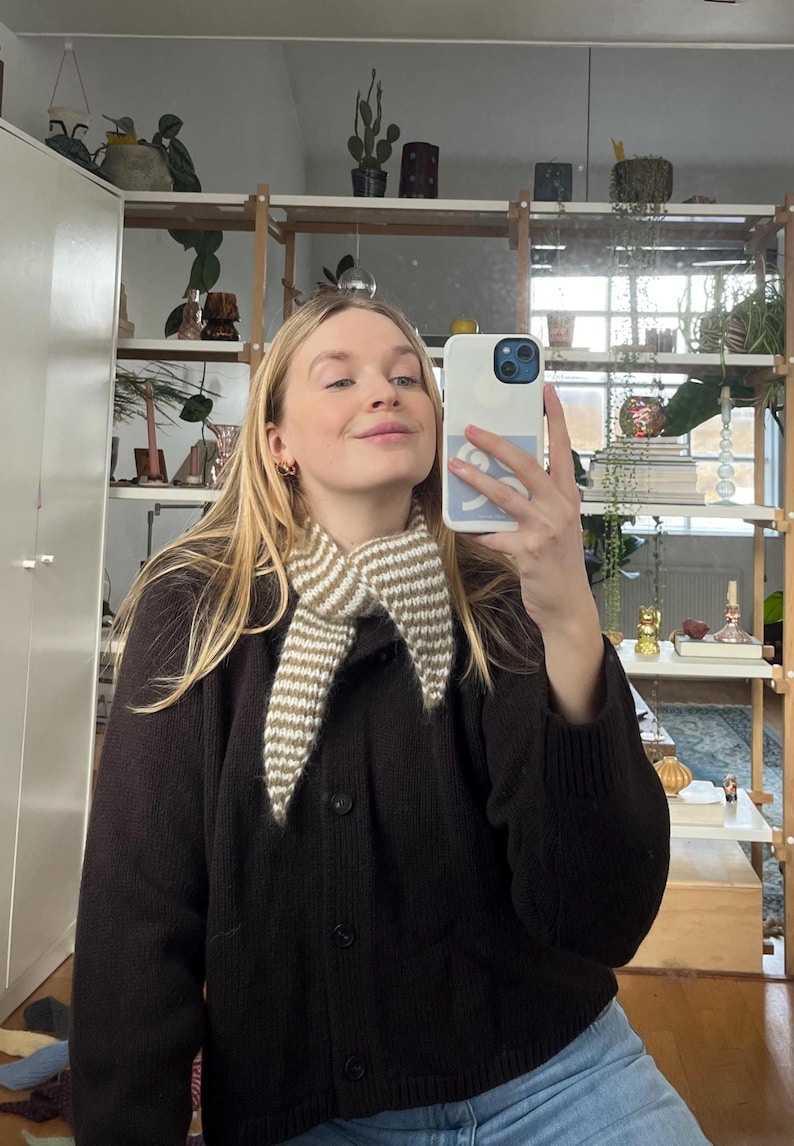 May include: A brown cardigan with a cream and brown striped collar. The cardigan is buttoned up and worn with blue jeans. The person is holding a white phone with a blue case and taking a selfie in a mirror.