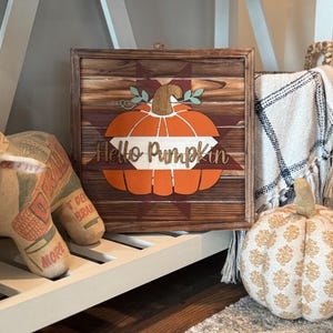 以下が含まれることがあります： オレンジ色のカボチャのデザインと「Hello Pumpkin」の文字が入った木製サイン。サインは濃い茶色のフレームと素朴な木材の背景を持っています。秋をテーマにした装飾品です。