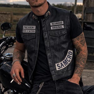 May include: Black leather vest with patches displaying "PRESIDENT MEN OF MAYHEM", "REDWOOD ORIGINAL", and "SAMCRO". The vest is worn over a black t-shirt and paired with black jeans. A motorcycle is visible in the background.