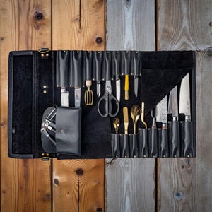 Pode incluir: Um rolo de facas de couro preto aberto, exibindo várias ferramentas de cozinha. O rolo contém facas, tesouras e outros utensílios, incluindo colheres douradas e uma pequena bolsa. O fundo é uma superfície de madeira com uma mistura de tábuas marrons e cinzas.