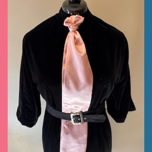May include: A black velvet dress with a high collar and three-quarter sleeves. A light pink satin panel runs down the front, tied at the neck. A black belt with a silver buckle cinches the waist.