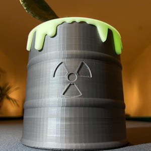 Könnte beinhalten: Ein schwarzer, zylindrischer Pflanztopf in Form eines radioaktiven Abfallbehälters mit einem grünen, tropfenden Schleimrand. Der Topf hat ein erhabenes, geprägtes Strahlungssymbol auf der Vorderseite.