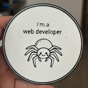 May include: A circular white coaster with a black border featuring a black line drawing of a smiling spider. Above the spider, the text reads "I'm a web developer" in a simple black sans-serif font. The design is centred on the white surface.