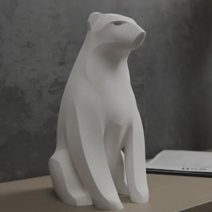 May include: A matte white sculpture of a polar bear in a seated position. The figure features smooth, angular, and geometric surfaces that define the bear's head, neck, and body, with a forward-facing gaze and a clean, solid, and uniform finish.