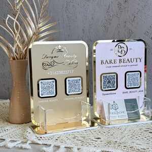 以下が含まれることがあります： 「Daryna Beauty」と「Bare Beauty」の文字が入った、2つのゴールドカラーのアクリル製名刺ホルダー。各ホルダーにはQRコードと、名刺用の透明アクリルポケットが付いています。背景には、装飾的な葉が入った花瓶があります。