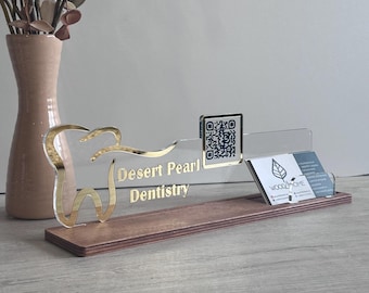 May include: A clear acrylic sign with a gold-coloured tooth icon and the text Desert Pearl Dentistry, featuring a gold-framed QR code and a slot holding business cards, all mounted on a rectangular wooden base with a smooth, dark finish.