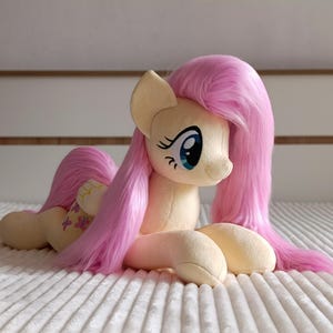 May include: A plush toy pony with a light yellow body, pink mane and tail, and blue eyes. The pony is lying down on a white, textured surface. The pony's tail has a floral design.