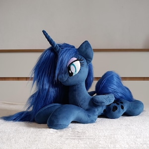 Könnte beinhalten: Ein dunkelblaues Plüschtier von Prinzessin Luna aus My Little Pony. Das Pony hat eine lange, fließende Mähne und einen Schweif. Es hat ein spiralförmiges Horn und große Augen mit Wimpern. Das Plüschtier liegt auf einer weißen Oberfläche.