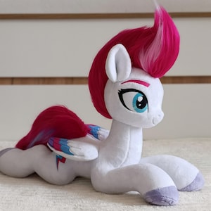 May include: A plush toy of a white pony with a bright pink mane and tail. The pony has large blue eyes, light purple hooves, and wings featuring blue, pink, and white accents. A small red lightning bolt symbol is visible on the pony's flank.