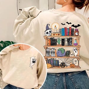 May include: A beige crewneck sweatshirt featuring a small ghost reading a book on the front left chest and a large graphic on the back depicting a three-tiered wooden bookshelf filled with books, skulls, a candle, a potion, and a spiderweb.