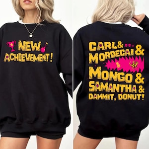 May include: Black sweatshirt with the text "NEW ACHIEVEMENT!" in yellow and pink pixelated font on the front. The back features the text "CARL & MORDECAI & MONGO & SAMANTHA & DAMMIT, DONUT!" in a similar style.