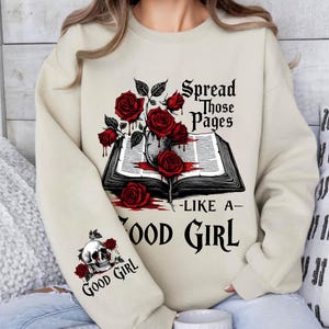 May include: Beige sweatshirt featuring a graphic of an open book with red roses and the text "Spread Those Pages Like a Good Girl." The left sleeve displays a skull with roses and the words "Good Girl."