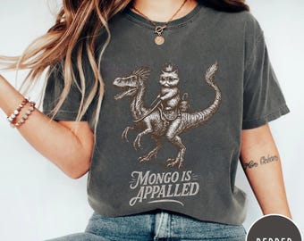 May include: Dark grey t-shirt featuring a cat riding a dinosaur with the text "MONGO IS APPALLED" in a vintage style. The graphic is in brown tones. The shirt is a Pepper color.