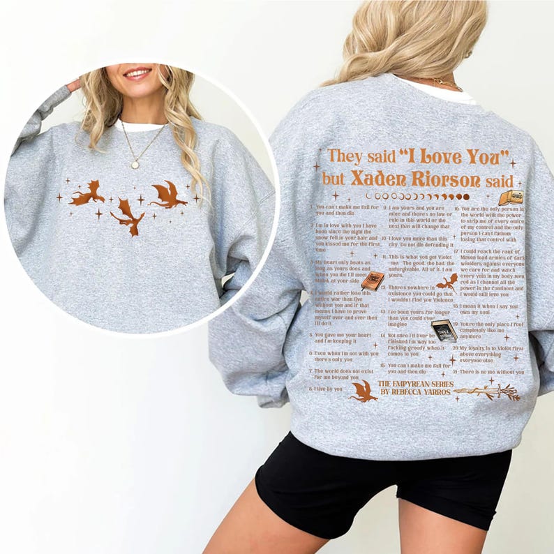 May include: A light gray sweatshirt with a white collar featuring a dragon design and text. The front displays three brown dragons and stars. The back has text that reads "They said 'I Love You' but Xaden Riorson said..." and "The Empyrean Series by Rebecca Yarros."