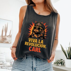 May include: A charcoal grey vest top with a cartoon cat wearing sunglasses and a beret, set against a fiery background. The text "VIVA LA REVOLUCIÓN, CARL" is printed in yellow and red.