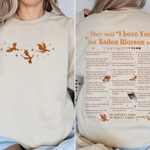 May include: Beige sweatshirt featuring a dragon graphic and text. The front displays three orange dragons and stars. The back has the text "They said 'I Love You' but Xaden Riorson said" with a list of quotes from the book series. The Empyrean Series by Rebecca Yarros.