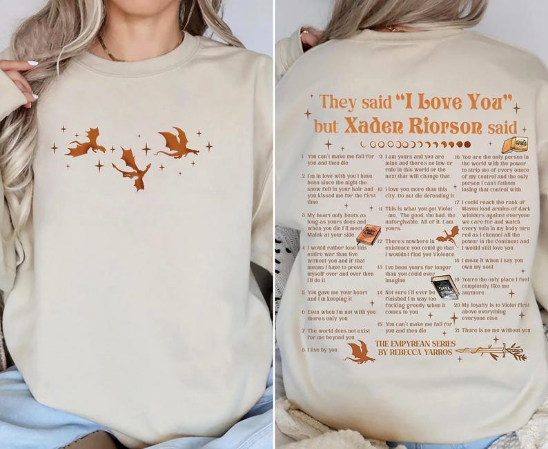 May include: Beige sweatshirt featuring a dragon graphic and text. The front displays three orange dragons and stars. The back has the text "They said 'I Love You' but Xaden Riorson said" with a list of quotes from the book series. The Empyrean Series by Rebecca Yarros.