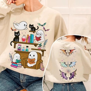 Könnte beinhalten: Ein beigefarbenes Sweatshirt mit Rundhalsausschnitt und einem Frontdruck von Geistern und schwarzen Katzen, die Bücher in Regalen neben Zaubertrankflaschen lesen. Auf der Rückseite sind drei Sätze bunter Fledermausflügel mit feinen Linien abgebildet.
