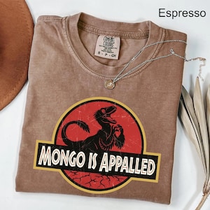 May include: Espresso-colored t-shirt with a graphic featuring a black dinosaur silhouette inside a red circle with the text "MONGO IS APPALLED" in white. The shirt has a crew neck and a small tag. A silver necklace with a pendant is on the shirt.