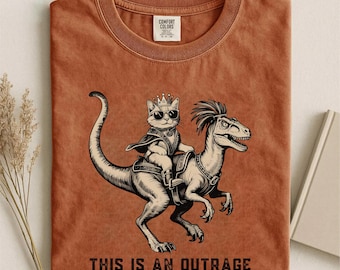 May include: Rust-colored t-shirt featuring a graphic of a cat wearing sunglasses and a crown, riding a dinosaur. The text "THIS IS AN OUTRAGE" is printed below the image. The shirt is made of a soft material.