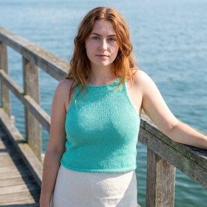 May include: A turquoise knitted halter top. The top is sleeveless and has a high neckline. The person is standing on a wooden pier with the ocean in the background. The top is paired with a white skirt.