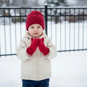 Op de afbeelding: Een kind met een rode gebreide muts en bijpassende wanten staat in de sneeuw. Het kind draagt ook een beige jas en een blauwe broek. Op de achtergrond is een zwart hek te zien.